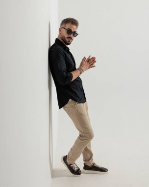 Güneş gözlüklü, kendine güvenen, avuçlarını ovuşturan ve arka planda gri bir duvara yaslanan havalı bir adam.