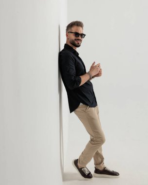 Güneş gözlüklü, siyah gömlekli mutlu bir genç, duvara yaslanmış, parmaklarına dokunuyor ve gri arka planda bir duvara yaslanıyor.