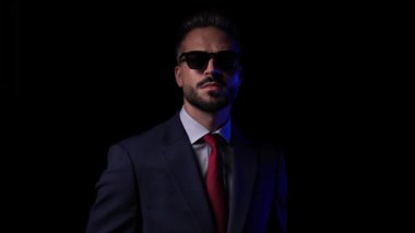 Güneş gözlüklü zarif iş adamı, siyah arkaplan önünde lacivert takım elbise ve güneş gözlüğünü ayarlamaya çalışıyor.