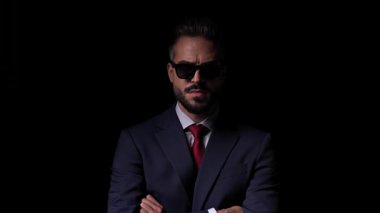 Güneş gözlüklü yakışıklı adam kollarını kavuşturup siyah arka planın önüne bakıyor.