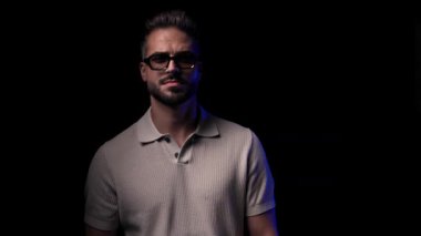 Güneş gözlüklü, kollarını kavuşturup siyah arka planın önünde yürüyen mutlu bir adam.