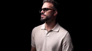 Güneş gözlüklü, gülümseyen, sakala dokunan ve siyah arka planda kollarını kavuşturan havalı genç adamın yan görüntüsü.