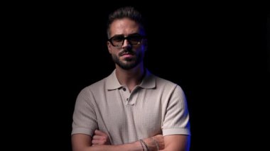 Güneş gözlüklü havalı adam kollarını kavuşturuyor, çenesine dokunuyor, yana bakıyor ve siyah arka planın önünde duruyor.