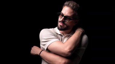 Günlük sakallı, güneş gözlüklü, kendine sarılan, gülümseyen ve siyah arka planda yürüyen bir adamın videosu.