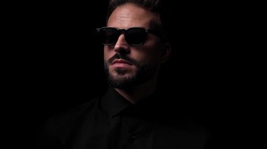 Siyah gözlüklü, güneş gözlüğü takan, güneş gözlüğünü ayarlayan ve siyah arka planda yürüyen seksi genç adam.