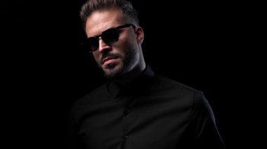 Havalı moda adamı poz veriyor ve güneş gözlüğünü ayarlıyor, siyah arka planın önünde duruyor ve yan tarafa bakıyor.