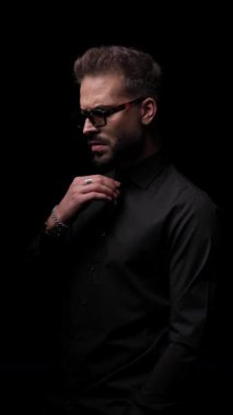 Güneş gözlüklü dramatik bir modacı gömleğini ayarlamaya çalışıyor, el ele tutuşuyor, avuç içlerini ovuyor ve siyah arka planda poz veriyor.