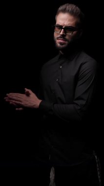 Avuçlarını ovuşturan, ellerini cebine sokan ve siyah arka plan arayan havalı moda adamının yan görüntüsü.