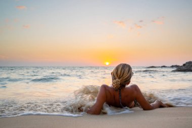 Gün batımında kumlu bir okyanusta mayo giymiş güzel bir kadın.