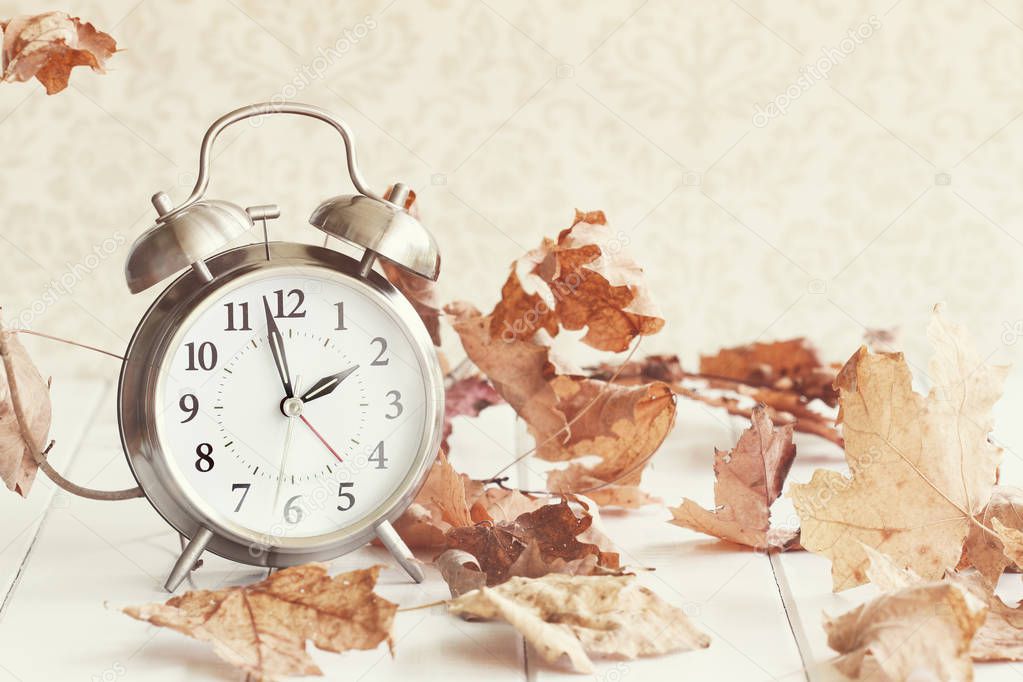 Faded Alarm clock in colorful autumn leaves against a retro background with shallow depth of field. Daylight savings time concept.