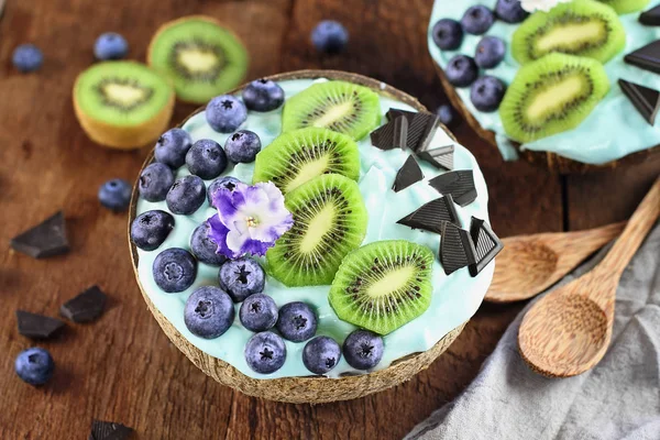 蓝色螺旋藻和浆果冰沙碗 新鲜的蓝莓 猕猴桃和巧克力片与木制勺子在椰子碗在质朴的背景 从上面 头顶拍摄的图像 — 图库照片