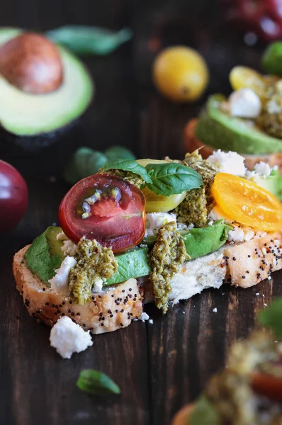 Avocado toast sandwich with avocados, pesto, fetaa cheese, fresh from the garden basil and heirloom tomatoes, over a rustic wooden background. Greek food and healthy vegetarian diet concept.