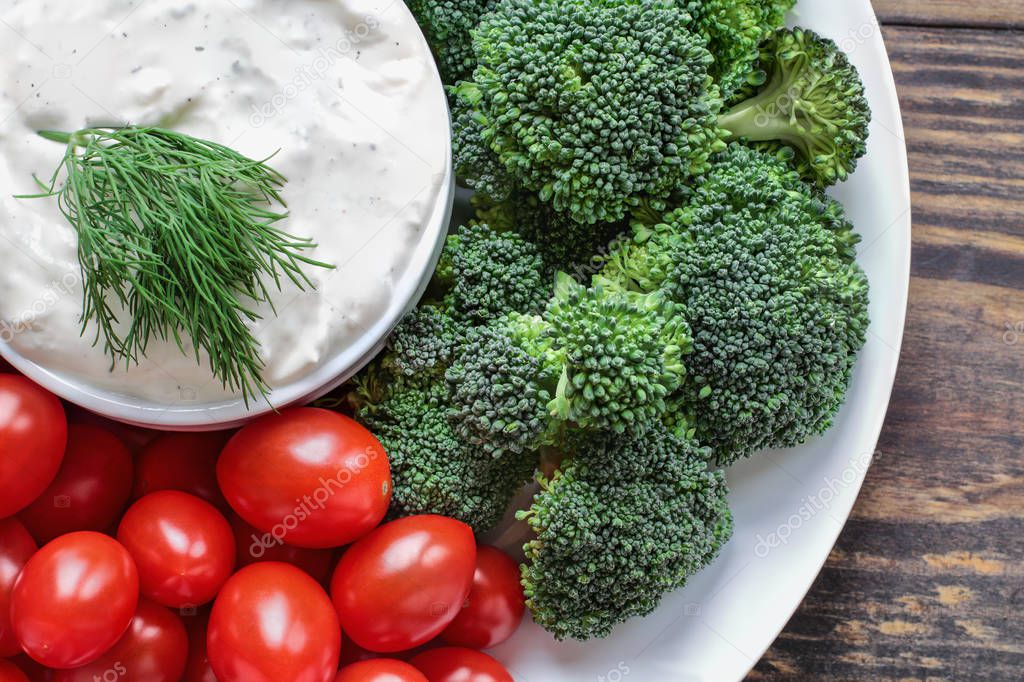 Homemade buttermilk ranch salad dressing with dill served with fresh cherry tomatoes and broccoli over a rustic wooden background. Image shot above from top view.