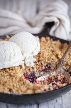 Böğürtlenli ve yaban mersinli tart dökme demir bir tavada pişmiş ve üzerinde de kırsal beyaz ahşap bir masanın üzerinde dondurmalı altın yulaf ezmesi çıtırı. Bulanık arkaplan ile aşırı seçici odak.