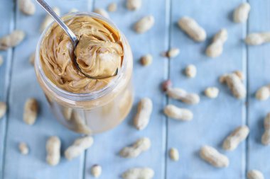 Fıstık ezmesi, kabuğunda fındık parçaları olan mavi bir masanın üzerinde plastik bir kavanozdan kaşıkla alınıyor. Bulanık arkaplan ile seçmeli odak. 