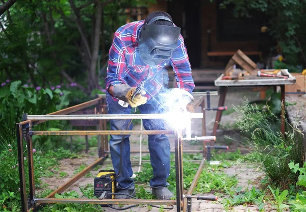 Homem em uma máscara protetora soldagem construção de metal fora da casa em um dia de verão — Fotografia de Stock