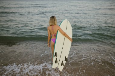 Sörf tahtası taşıyan bir kadın güneşli bir sahilde dalgalara doğru yürür, sörf seansına hazırdır. Bu arka plan onun okyanusla olan bağlantısını vurguluyor ve yaz temaları için mükemmel..
