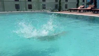 Çocuk güneşli bir yaz gününde havuza atlar, havada yakalanır. Temiz mavi su, sahil oteli, eğlenceli tatil aktiviteleri.