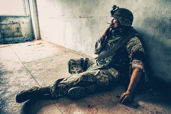 戦闘服の兵士と弾薬ベルトの胸の上 床に座って 後喫煙が肩で撮影し 周りの落とした武器の血で近くにある汚れの負傷者の手の包帯を置く — ストック写真