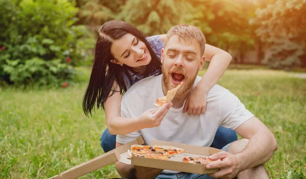 Young couple eating pizza in the park. They treat each other and laugh.