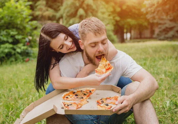 Young couple eating pizza in the park. They treat each other and laugh.