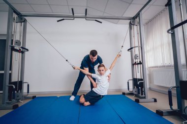 Rehabilitasyon kavramı. Genç çocuk mat fizyoterapist gözetiminde egzersizleri yapıyor. Egzersiz tedavisi