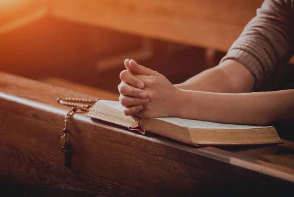 Christian woman praying in church. Hands crossed and Holy Bible on wooden desk. Background