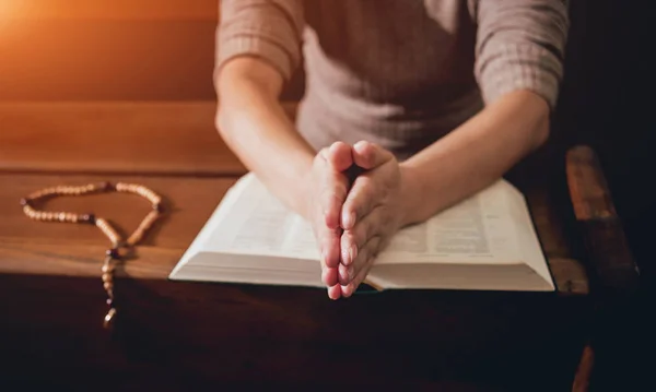 Christian woman praying in church. Hands crossed and Holy Bible on wooden desk. Background