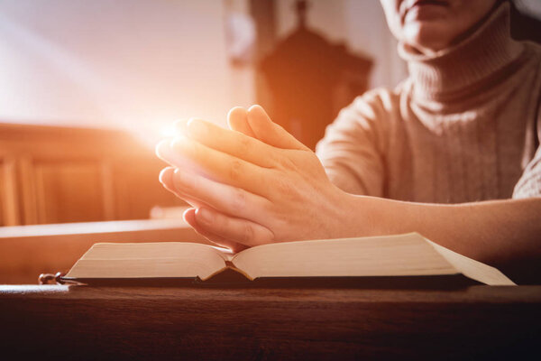 Christian woman praying in church. Hands crossed and Holy Bible on wooden desk. Background