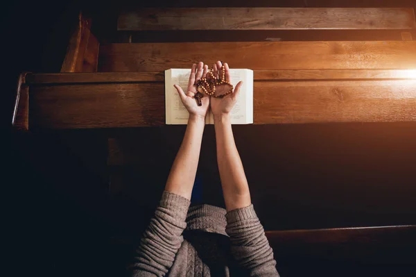 Christian woman praying in church. Hands crossed and Holy Bible on wooden desk. Background