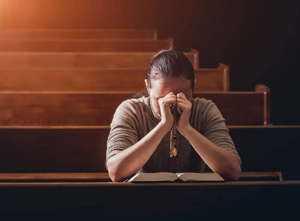 Christian woman praying in church. Hands crossed and Holy Bible on wooden desk. Background