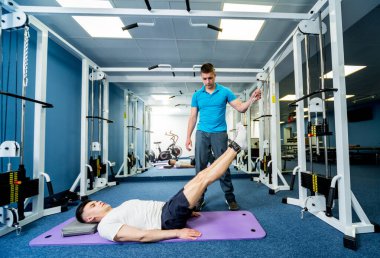 Rehabilitasyon terapisi. Fizyoterapistin gözetiminde paspas üzerinde egzersiz yapan genç bir adam. Omurgada tedavi ağrısı