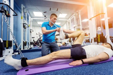 Rehabilitasyon terapisi. Fizyoterapistin gözetiminde paspas üzerinde egzersiz yapan genç bir adam. Omurgada tedavi ağrısı