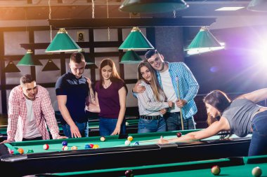 Bilardo oynayan genç bir kadın. Elinde ıstakayla masanın yanında poz veriyordu. Arka planda bir grup arkadaş.