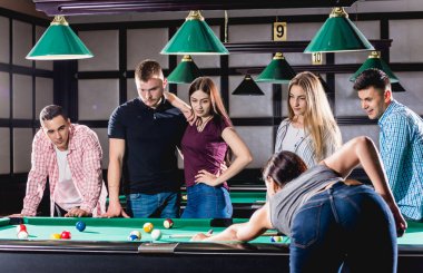 Bilardo oynayan genç bir kadın. Elinde ıstakayla masanın yanında poz veriyordu. Arka planda bir grup arkadaş.