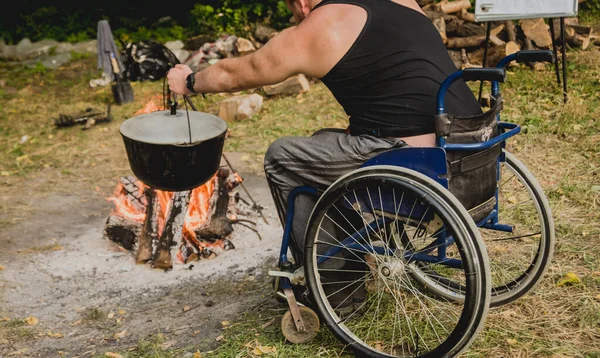 Disabled man resting in a campsite with friends. Wheelchair in the forest on the background of bonfire. Barbeque. Camping. Summertime