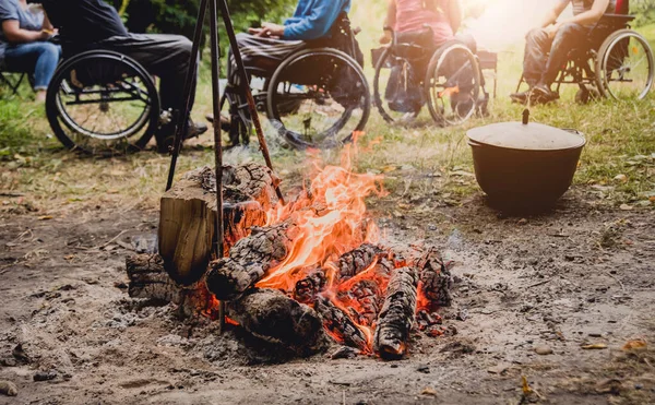 Group of disabled persons resting in a campsite with friends. Wheelchair in the forest on the background of bonfire. Barbeque. Camping. Summertime