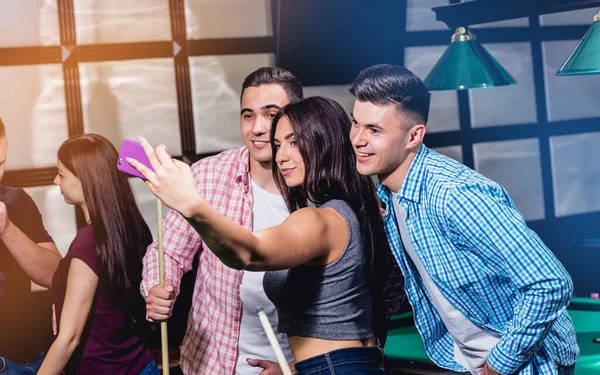 Un grupo de amigos hace una selfie en la mesa de billar. Posando con un taco en sus manos . — Foto de Stock