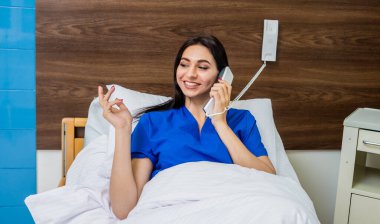 Hasta hastane yatağında telefonla konuşuyor. Genç, beyaz bir kadın. Hemşireye bağlı