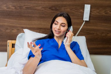 Hasta hastane yatağında telefonla konuşuyor. Genç, beyaz bir kadın. Hemşireye bağlı