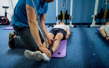 Rehabilitasyon terapisi. Fizyoterapistin gözetiminde paspas üzerinde egzersiz yapan genç bir adam. Omurgada tedavi ağrısı