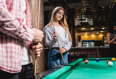 Bilardo oynayan genç bir kadın. Elinde ıstakayla masanın yanında poz veriyordu. Arka planda bir grup arkadaş.