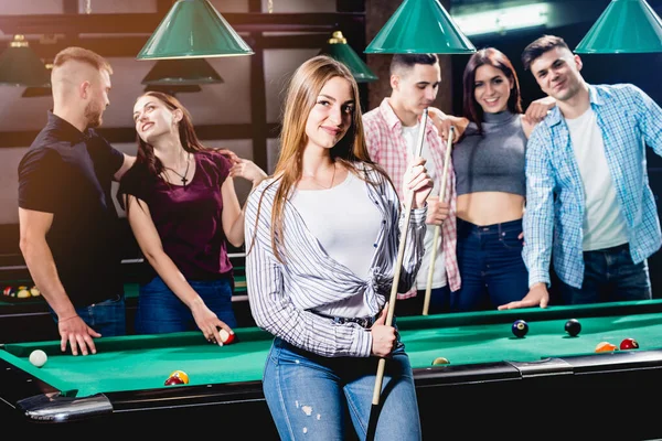 Young woman playing in billiard. Posing near the table with a cue in her hands. A group of friends on the background.
