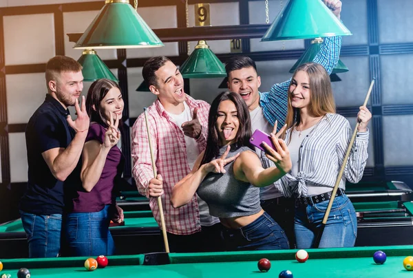 A group of friends makes a selfie at the pool table. Posing with a cue in their hands.
