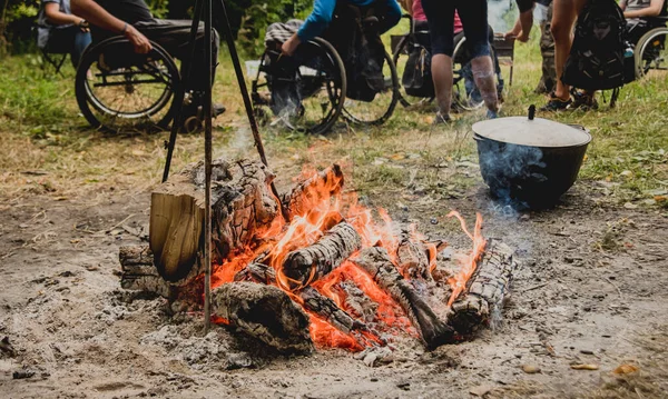 Group of disabled persons resting in a campsite with friends. Wheelchair in the forest on the background of bonfire. Barbeque. Camping. Summertime