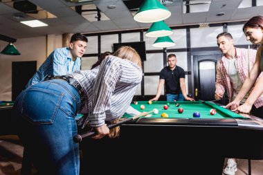 Bilardo oynayan genç bir kadın. Elinde ıstakayla masanın yanında poz veriyordu. Arka planda bir grup arkadaş.