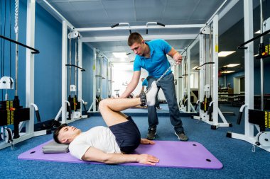 Rehabilitasyon terapisi. Fizyoterapistin gözetiminde paspas üzerinde egzersiz yapan genç bir adam. Omurgada tedavi ağrısı