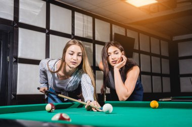 Bilardo oynayan iki genç kadın. Arka planda bilardo odası..