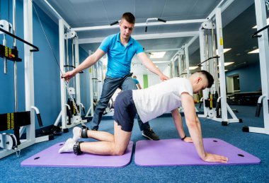 Rehabilitasyon terapisi. Fizyoterapistin gözetiminde paspas üzerinde egzersiz yapan genç bir adam. Omurgada tedavi ağrısı