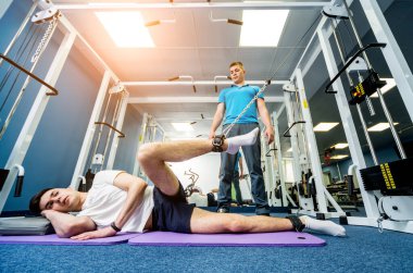 Rehabilitasyon terapisi. Fizyoterapistin gözetiminde paspas üzerinde egzersiz yapan genç bir adam. Omurgada tedavi ağrısı